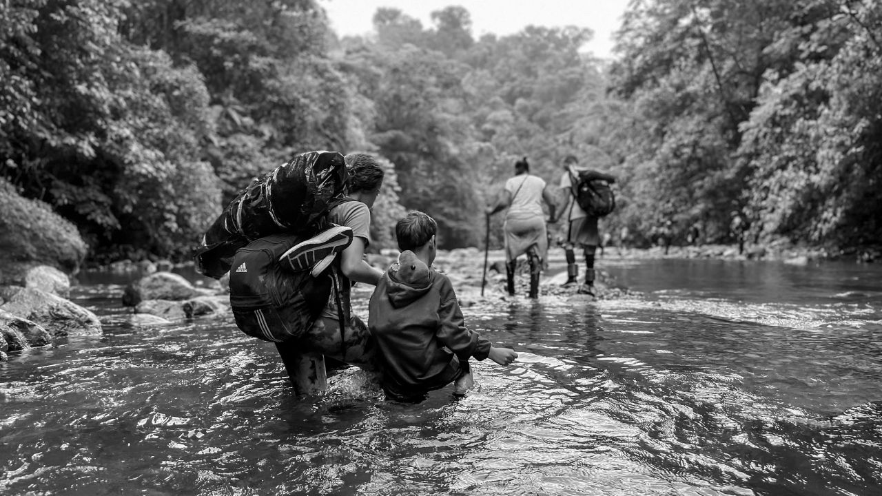 Une femme vénézuélienne guide son fils dans l’eau jusqu’aux genoux, son jouet de dinosaure fourré en toute sécurité dans son sweat-shirt. Elle espère les amener aux États-Unis, pour retrouver son mari.